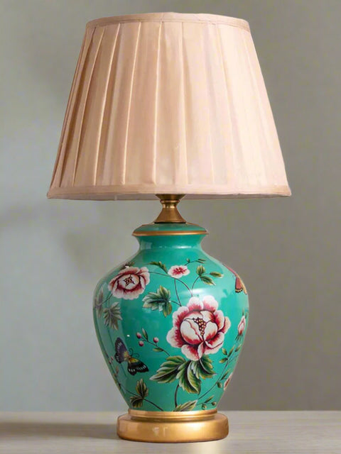 Tatva Lights Bloom Ceramic Table Lamp featuring a vibrant turquoise base adorned with hand-painted floral and butterfly motifs, complemented by gold accents and a pleated beige lampshade—an elegant fusion of color and charm for classic or eclectic interiors.