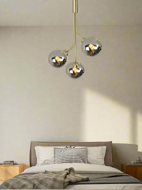 A modern style pendant ceiling light with three LED lights, featuring a gold-colored fixture and clear glass shades, installed over a bed in a bedroom setting.