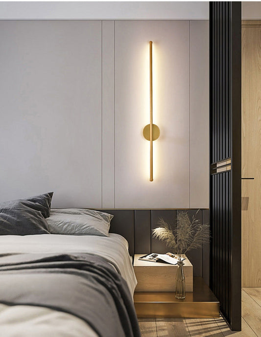 An 800mm long sleek tube wall light in glossy French gold, mounted on a wall in a bedroom setting, providing illumination.