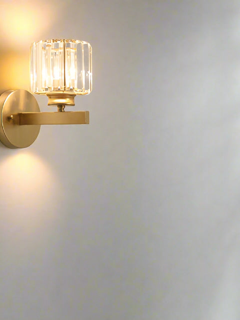 A gold crystal glass shade wall light mounted on a wall, with a luxurious living room in the background featuring a brown sofa and a side table with a clock.
