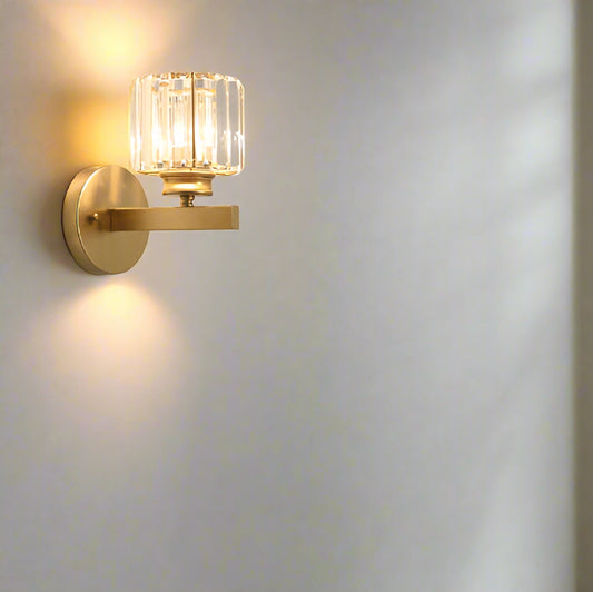 A gold crystal glass shade wall light mounted on a wall, with a luxurious living room in the background featuring a brown sofa and a side table with a clock.