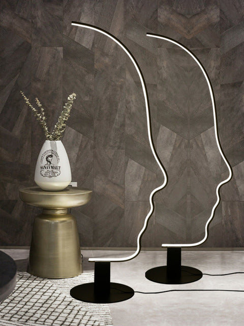 A black face arc floor standing lamp with a warm white light, placed on a floor next to a small round table with a vase of flowers, in a room with wooden flooring and a patterned wall.