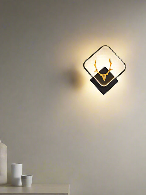A gold stag-shaped LED wall lamp mounted on a wall, with warm white lights, positioned above a brown armchair in a room with a gray wall and a television in the background.