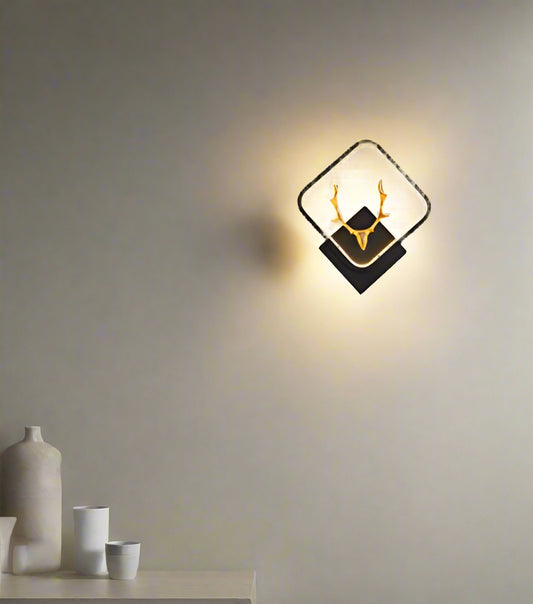 A gold stag-shaped LED wall lamp mounted on a wall, with warm white lights, positioned above a brown armchair in a room with a gray wall and a television in the background.