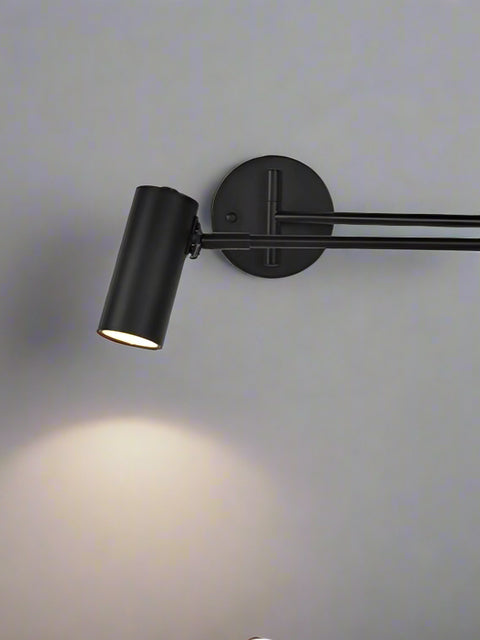Modern LED black long arm adjustable wall light mounted on a white wall above a white nightstand with a lamp on it, next to a bed with a green headboard.