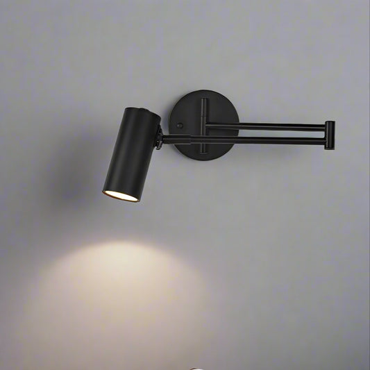 Modern LED black long arm adjustable wall light mounted on a white wall above a white nightstand with a lamp on it, next to a bed with a green headboard.