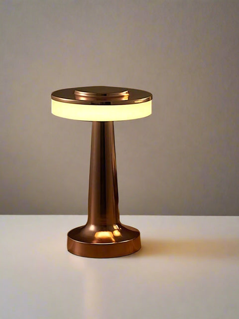 Tatva Lights MUSHED Touch Control Wireless Bar Table Lamp in a sleek copper finish, featuring a mushroom-inspired design with a circular light diffuser and a tapered base. Placed on a white tabletop with a magazine beside it, the lamp casts a warm ambient glow against a neutral wall.