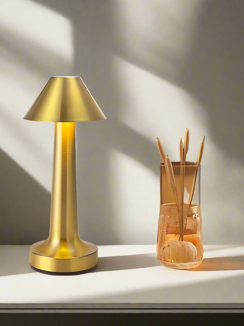 Tatva Lights MetalHat Touch-Control Rechargeable Table Lamp in a luxurious gold finish, featuring a sleek conical hat-shaped shade and slender stem. Positioned on a modern white bedside table with decorative elements, this elegant lamp offers a warm ambient glow—ideal for contemporary bedroom or living room decor.