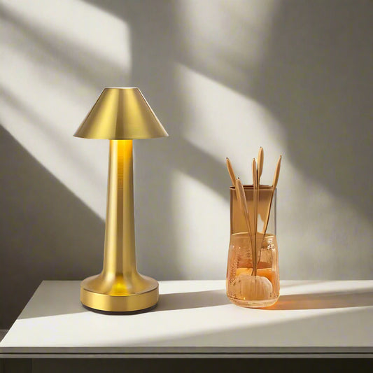 Tatva Lights MetalHat Touch-Control Rechargeable Table Lamp in a luxurious gold finish, featuring a sleek conical hat-shaped shade and slender stem. Positioned on a modern white bedside table with decorative elements, this elegant lamp offers a warm ambient glow—ideal for contemporary bedroom or living room decor.
