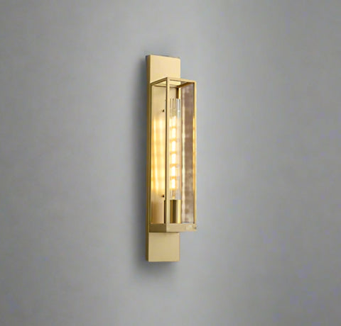 Modern gold metal wall light with a long transparent glass shade, mounted on a wall with stairs leading to it. The light emits a warm white glow.
