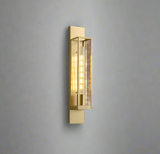 Modern gold metal wall light with a long transparent glass shade, mounted on a wall with stairs leading to it. The light emits a warm white glow.