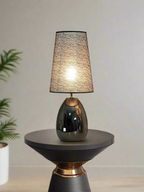 Tatva Lights Noir Table Lamp featuring a sleek, glossy black oval base and a textured cone-shaped shade, elegantly placed on a modern side table in a contemporary living room setting with muted tones and indoor greenery.