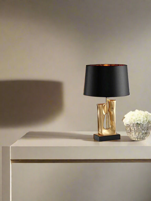 Tatva Lights' modern decorative table lamp featuring a sculpted gold base and black lampshade, styled on a sleek white console table beside a crystal vase of white flowers in a minimalist interior setting.
