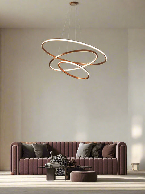 A modern rose gold LED chandelier with three concentric rings hanging over a stylish living room setting with a pink sofa and decorative wall art.