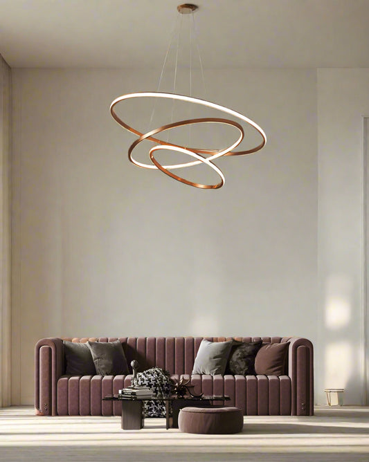 A modern rose gold LED chandelier with three concentric rings hanging over a stylish living room setting with a pink sofa and decorative wall art.