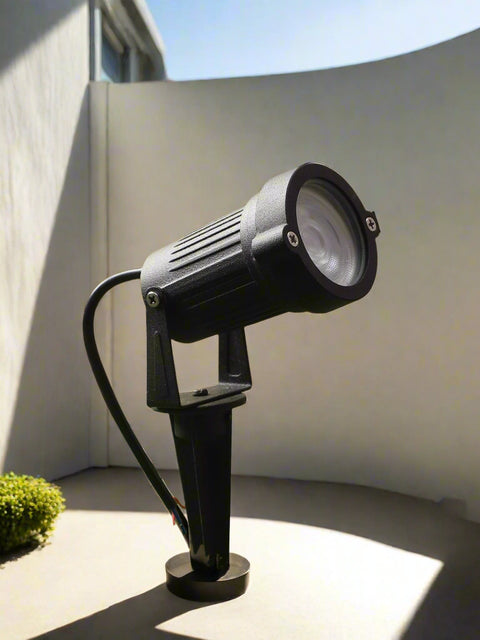 Tatva Lights black LED spike spotlight designed for outdoor use, ideal for illuminating landscapes, pathways, patios, architectural features, or garden areas, installed on artificial grass with warm ambient lighting and decorative plants in the background.