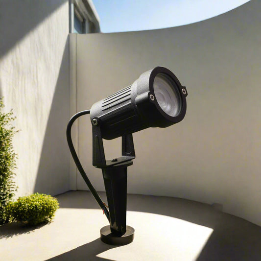 Tatva Lights black LED spike spotlight designed for outdoor use, ideal for illuminating landscapes, pathways, patios, architectural features, or garden areas, installed on artificial grass with warm ambient lighting and decorative plants in the background.