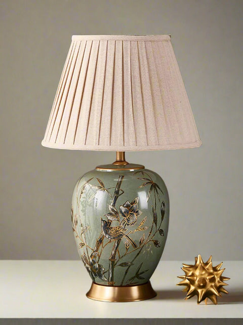 Tatva Lights Verdant Song ceramic table lamp featuring a hand-painted floral and bird motif on a soft green base with a golden pedestal, topped with a pleated beige fabric lampshade — perfect for elegant bedside or living room decor.