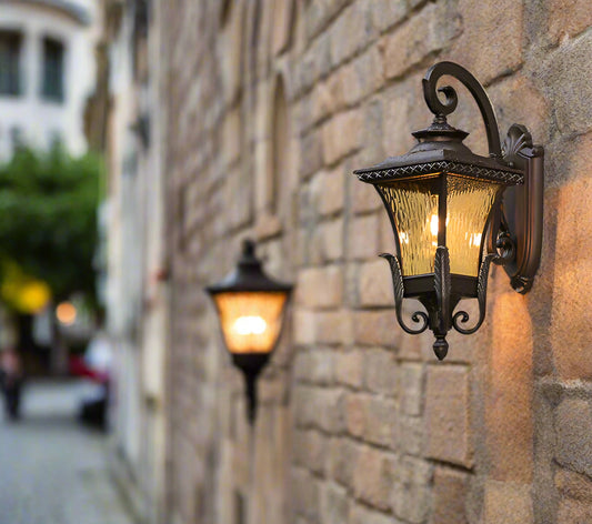 Tatva Lights black Victorian-style wall-mounted outdoor lantern featuring clear textured glass panels, elegantly installed beside a window on a beige exterior wall, casting a warm ambient glow.