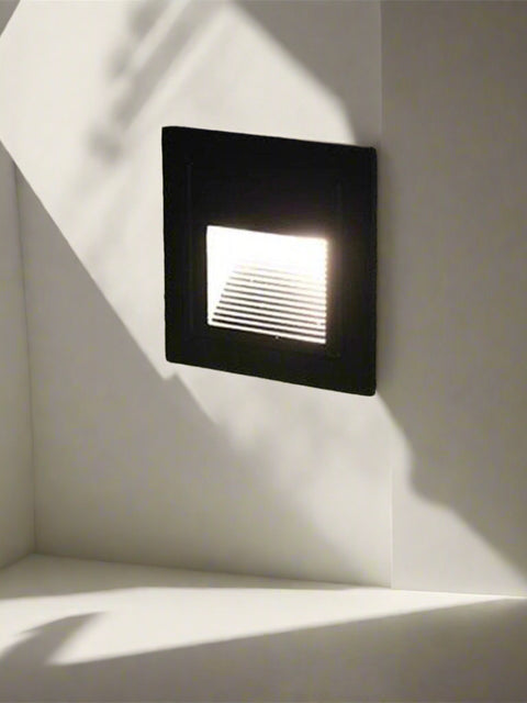 Black square recessed LED outdoor wall light installed on a textured wall, softly illuminating a garden pathway with focused, downward-facing light — ideal for steps, driveways, and landscape accents.