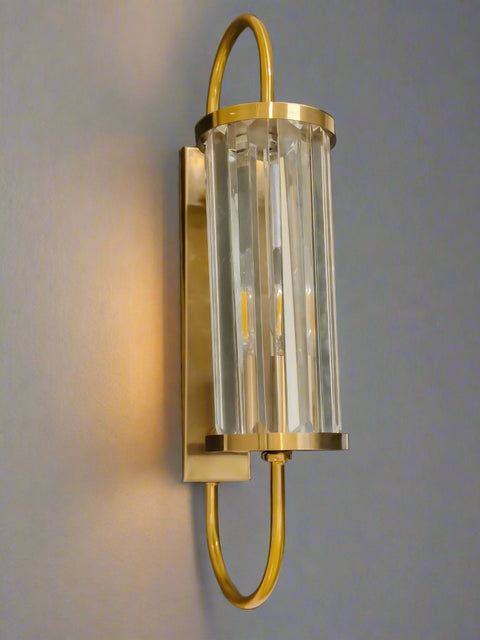 Tatva Lights Brionyx Gold Wall Sconce – elegant wall-mounted fixture featuring vertical glass prisms and a brushed gold frame with a curved design. Ideal for modern luxury interiors, hallways, and accent walls seeking warm, ambient lighting with a sophisticated touch.
