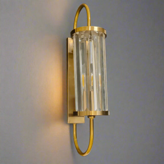Tatva Lights Brionyx Gold Wall Sconce – elegant wall-mounted fixture featuring vertical glass prisms and a brushed gold frame with a curved design. Ideal for modern luxury interiors, hallways, and accent walls seeking warm, ambient lighting with a sophisticated touch.
