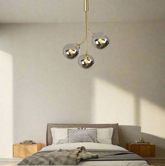 A modern style pendant ceiling light with three LED lights, featuring a gold-colored fixture and clear glass shades, installed over a bed in a bedroom setting.
