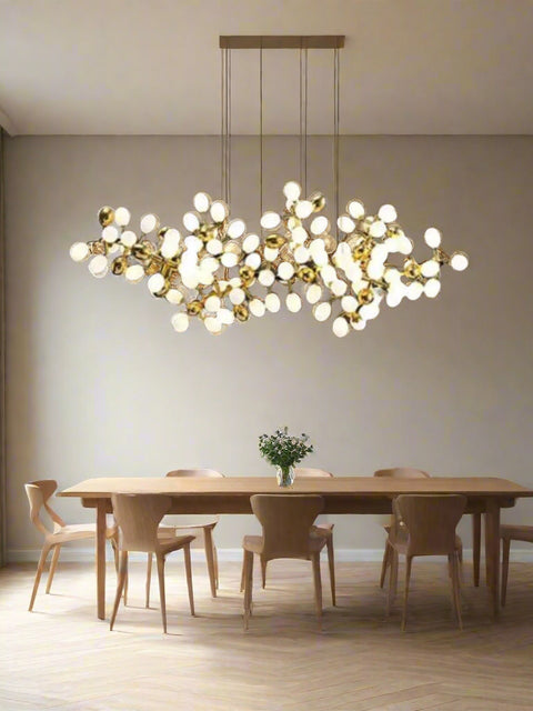 Tatva Lights Elegance Horizon Modern Chandelier suspended above a contemporary wooden dining table, featuring a sculptural arrangement of gold-finished branches with spherical frosted bulbs. The chandelier adds a luxurious, artistic touch to the minimalist dining room setting with natural light and neutral tones.