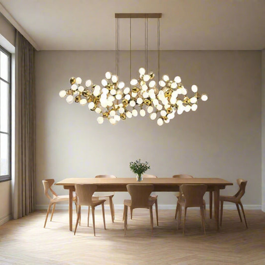 Tatva Lights Elegance Horizon Modern Chandelier suspended above a contemporary wooden dining table, featuring a sculptural arrangement of gold-finished branches with spherical frosted bulbs. The chandelier adds a luxurious, artistic touch to the minimalist dining room setting with natural light and neutral tones.