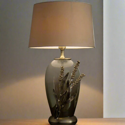 Tatva Lights Exquisite Handcrafted Table Lamp featuring a smoky glass base adorned with intricate metallic floral detailing and topped with a soft taupe fabric lampshade—an artfully crafted piece that brings warmth, elegance, and natural charm to any space.