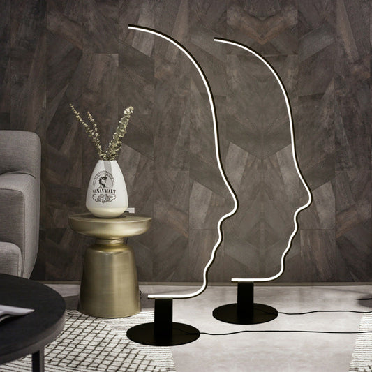 A black face arc floor standing lamp with a warm white light, placed on a floor next to a small round table with a vase of flowers, in a room with wooden flooring and a patterned wall.
