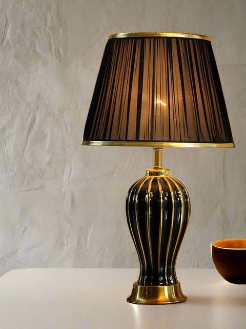 Tatva Lights Golden Accent Ceramic Table Lamp featuring a sleek black ceramic base with vertical gold accents and a matching black pleated lampshade with gold trim—an elegant lighting piece that adds warmth and luxury to any setting.