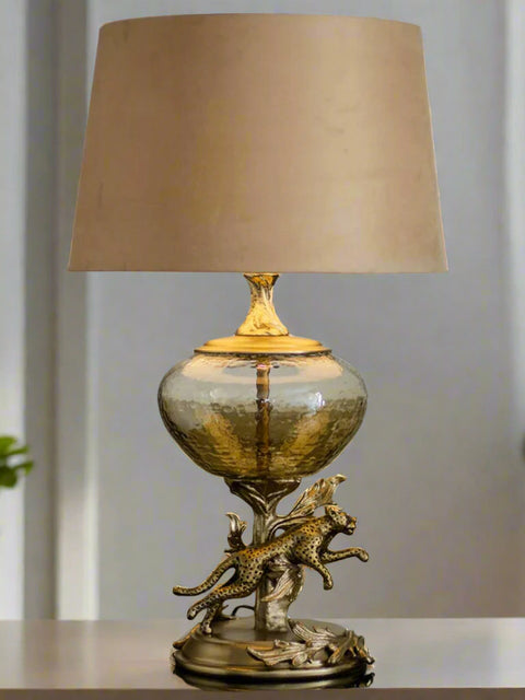 Tatva Lights Leopard Metal Glass Table Lamp featuring a hammered glass globe base with a sculpted golden leopard leaping from ornamental foliage, topped with a neutral beige lampshade for a bold and artistic home décor accent.