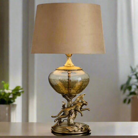 Tatva Lights Leopard Metal Glass Table Lamp featuring a hammered glass globe base with a sculpted golden leopard leaping from ornamental foliage, topped with a neutral beige lampshade for a bold and artistic home décor accent.