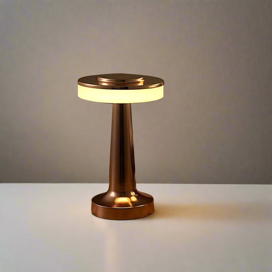 Tatva Lights MUSHED Touch Control Wireless Bar Table Lamp in a sleek copper finish, featuring a mushroom-inspired design with a circular light diffuser and a tapered base. Placed on a white tabletop with a magazine beside it, the lamp casts a warm ambient glow against a neutral wall.