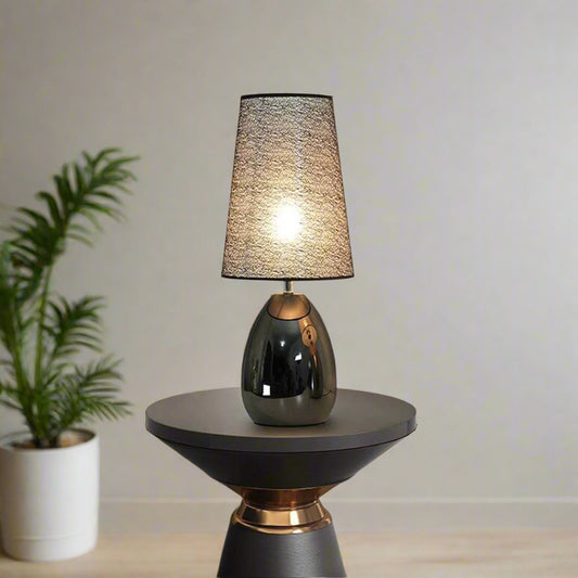 Tatva Lights Noir Table Lamp featuring a sleek, glossy black oval base and a textured cone-shaped shade, elegantly placed on a modern side table in a contemporary living room setting with muted tones and indoor greenery.