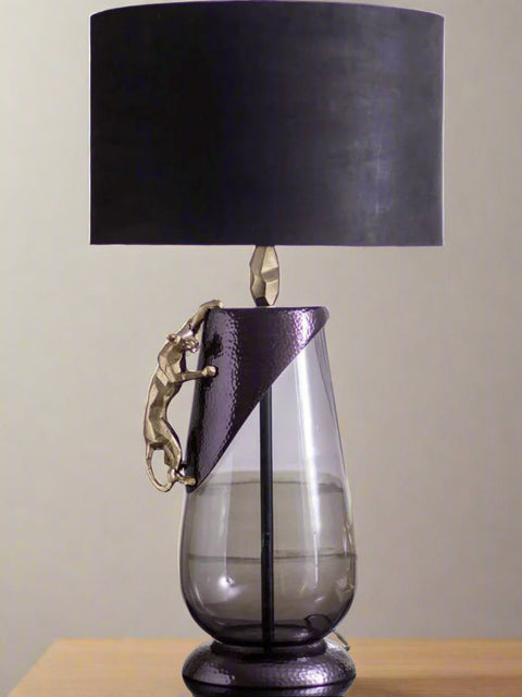 Tatva Lights Panther Metal Glass Table Lamp showcasing a sleek glass base with a textured metallic accent and a gold panther sculpture climbing its side, topped with a bold black lampshade for a dramatic and luxurious decor statement.
