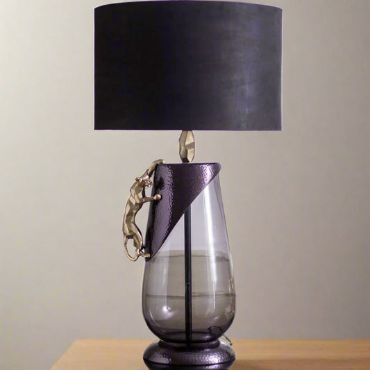 Tatva Lights Panther Metal Glass Table Lamp showcasing a sleek glass base with a textured metallic accent and a gold panther sculpture climbing its side, topped with a bold black lampshade for a dramatic and luxurious decor statement.