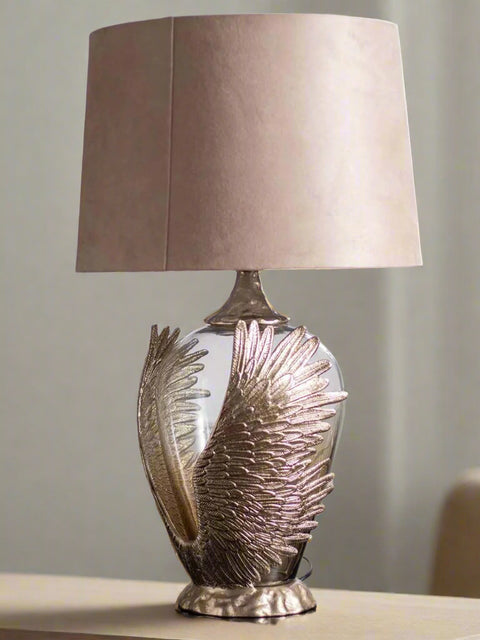 Tatva Lights Phoenix Wings Metal Glass Table Lamp featuring a sculpted golden wing-shaped base embracing a rounded glass body, paired with a neutral beige shade—ideal for luxurious and symbolic home décor styling.