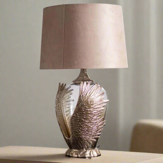Tatva Lights Phoenix Wings Metal Glass Table Lamp featuring a sculpted golden wing-shaped base embracing a rounded glass body, paired with a neutral beige shade—ideal for luxurious and symbolic home décor styling.