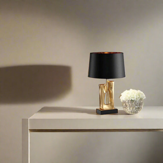 Tatva Lights' modern decorative table lamp featuring a sculpted gold base and black lampshade, styled on a sleek white console table beside a crystal vase of white flowers in a minimalist interior setting.