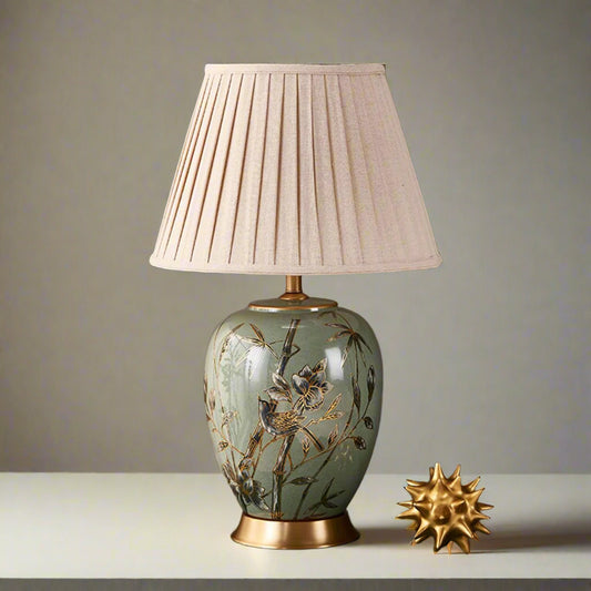 Tatva Lights Verdant Song ceramic table lamp featuring a hand-painted floral and bird motif on a soft green base with a golden pedestal, topped with a pleated beige fabric lampshade — perfect for elegant bedside or living room decor.