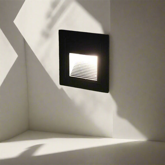 Black square recessed LED outdoor wall light installed on a textured wall, softly illuminating a garden pathway with focused, downward-facing light — ideal for steps, driveways, and landscape accents.